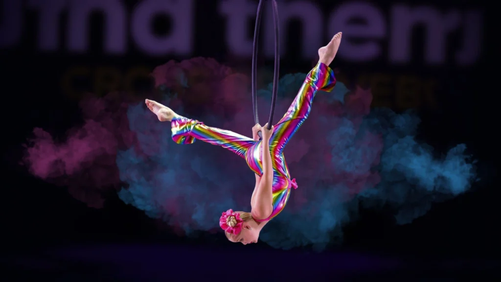 Female aerial performer in a rainbow jumpsuit hanging upside down on a hoop with colorful smoke effects in the background.