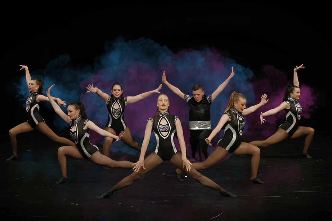 Group of seven competitive dancers in black and silver costumes performing a synchronized routine on stage with colorful smoke in the background.